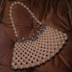 May include: A handbag constructed from interconnected white faux pearls, featuring a single pearl handle and a top border of faceted silver-toned beads. A small rectangular metal tag with the word OCEANS is attached to the front upper edge.