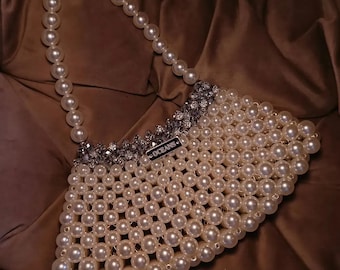Puede incluir: Un bolso hecho de perlas blancas artificiales interconectadas, con un asa de perlas y un borde superior de cuentas plateadas facetadas. Una pequeña etiqueta metálica rectangular con la palabra OCEANS está fijada en el borde superior frontal.