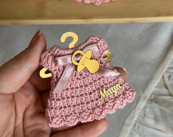 Puede incluir: Un pequeño adorno de vestido tejido a ganchillo en color rosa empolvado, colgado de una percha de metal dorado. El vestido está decorado con un lazo de satén rosa claro, un dije de chupete dorado y una placa dorada con el nombre Maya.