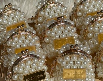 Puede incluir: Un conjunto de pequeños monederos cubiertos de perlas blancas, con marcos y cierres de metal dorado. Cada monedero tiene una pequeña etiqueta rectangular dorada con la fecha "05.29.2026". Los monederos están colocados sobre una bandeja de madera.