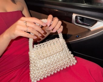 Puede incluir: Un bolso estructurado hecho completamente de perlas artificiales blancas interconectadas. El bolso presenta un cuerpo rectangular con cierre de solapa y un asa superior única, todo construido con cuentas de perlas esféricas, uniformes y brillantes.