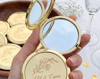 Puede incluir: Un espejo de bolsillo redondo de metal dorado con tapa abatible. El exterior presenta un diseño floral grabado y el texto "Elif & Caner WEDDING NIGHT 01 | 08 | 2026". El espejo está abierto, revelando dos superficies reflectantes en el marco.