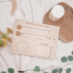 Op de afbeelding: Een lichtgekleurd houten babyspeelgoed met de tekst "MesattachestetinesAngel" en "Mes perles". Het heeft een cirkel van 7 cm, een sleuf van 22 cm en twee kleine cirkels. Een witte kaars en groen zijn op de achtergrond.