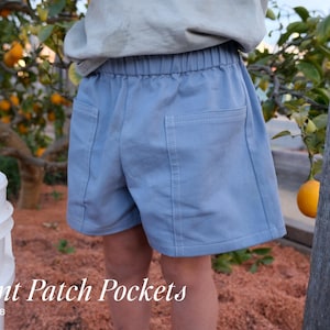 May include: Light blue shorts featuring an elastic waistband and two large square patch pockets on the front. The garment includes visible stitching details along the pocket edges. The text Front Patch Pockets VIEW A & B is displayed.