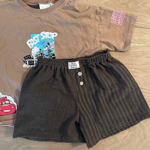 May include: A tan short-sleeved t-shirt featuring a graphic of a blue race car with the text FAST STOP and a red race car, paired with shorts that have a solid dark brown side and a dark brown and black striped side with two buttons.