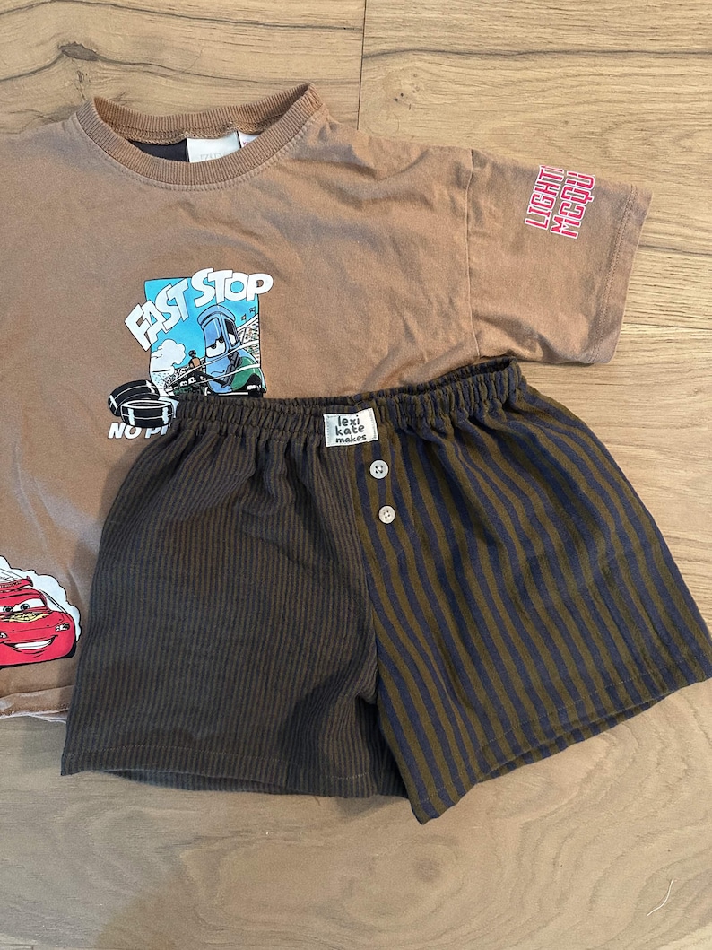 May include: A tan short-sleeved t-shirt featuring a graphic of a blue race car with the text FAST STOP and a red race car, paired with shorts that have a solid dark brown side and a dark brown and black striped side with two buttons.