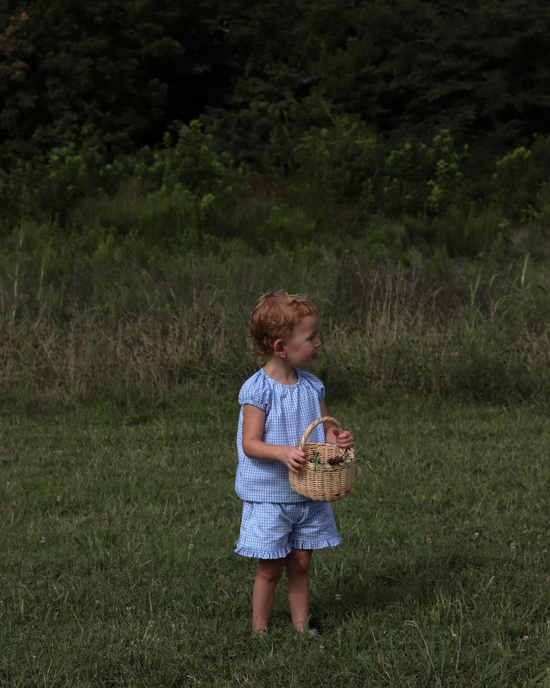 May include: A light blue and white gingham two-piece set consisting of a short-sleeved blouse with gathered shoulders and matching shorts with ruffled hems, paired with a small, round, light brown woven wicker basket with a handle.