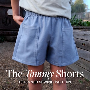 May include: Light blue shorts featuring an elastic waistband and two large patch pockets on the front. The text "The Tommy Shorts BEGINNER SEWING PATTERN" is printed in white at the bottom of the image over the fabric of the shorts.