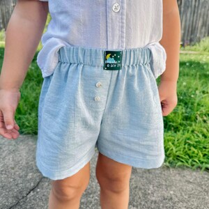 May include: Light blue shorts with an elastic waistband and two front buttons. A small dark green rectangular label is centered on the waistband featuring a yellow crescent moon, white stars, and the text "DREAM IT & DIY IT" in white.