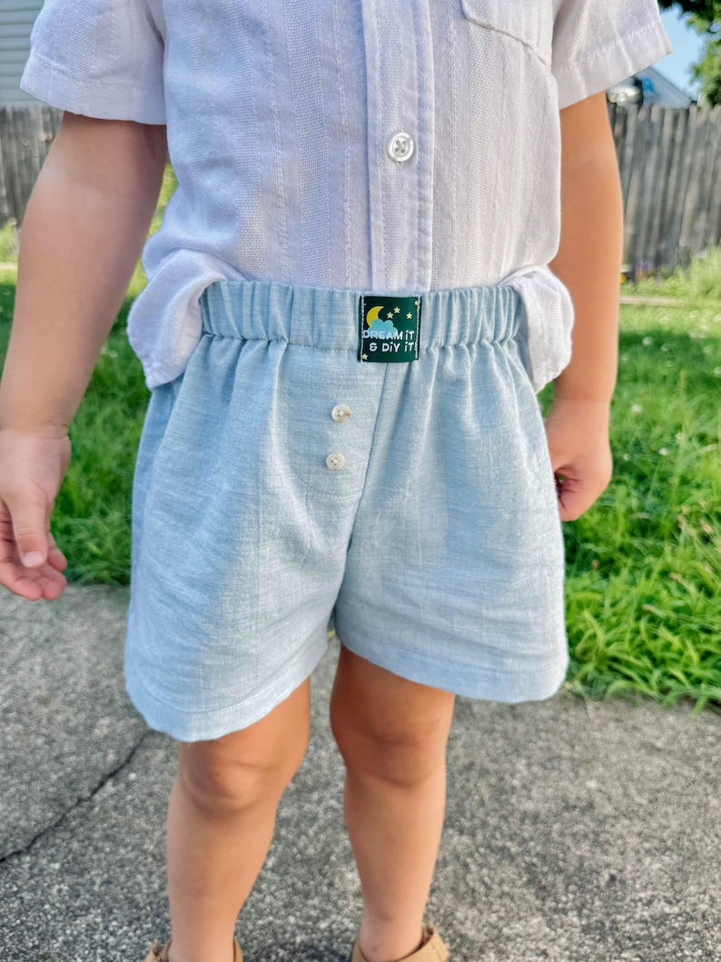 May include: Light blue shorts with an elastic waistband and two front buttons. A small dark green rectangular label is centered on the waistband featuring a yellow crescent moon, white stars, and the text "DREAM IT & DIY IT" in white.