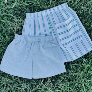 May include: Two pairs of shorts with elastic waistbands. One pair features a white fabric with vertical green stripes and a single patch pocket. The second pair is a solid light blue color with a textured appearance and a single patch pocket.