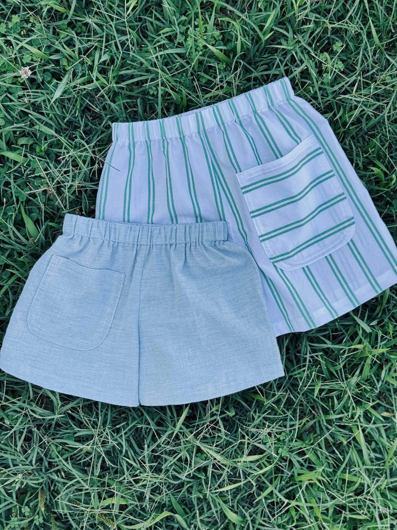 May include: Two pairs of shorts with elastic waistbands. One pair features a white fabric with vertical green stripes and a single patch pocket. The second pair is a solid light blue color with a textured appearance and a single patch pocket.