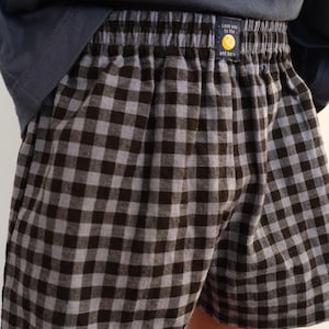May include: Boxer shorts featuring a black and gray gingham check pattern with an elastic waistband. A small rectangular label on the front center displays a yellow moon graphic and the text "Love you to the moon and back" in white font.