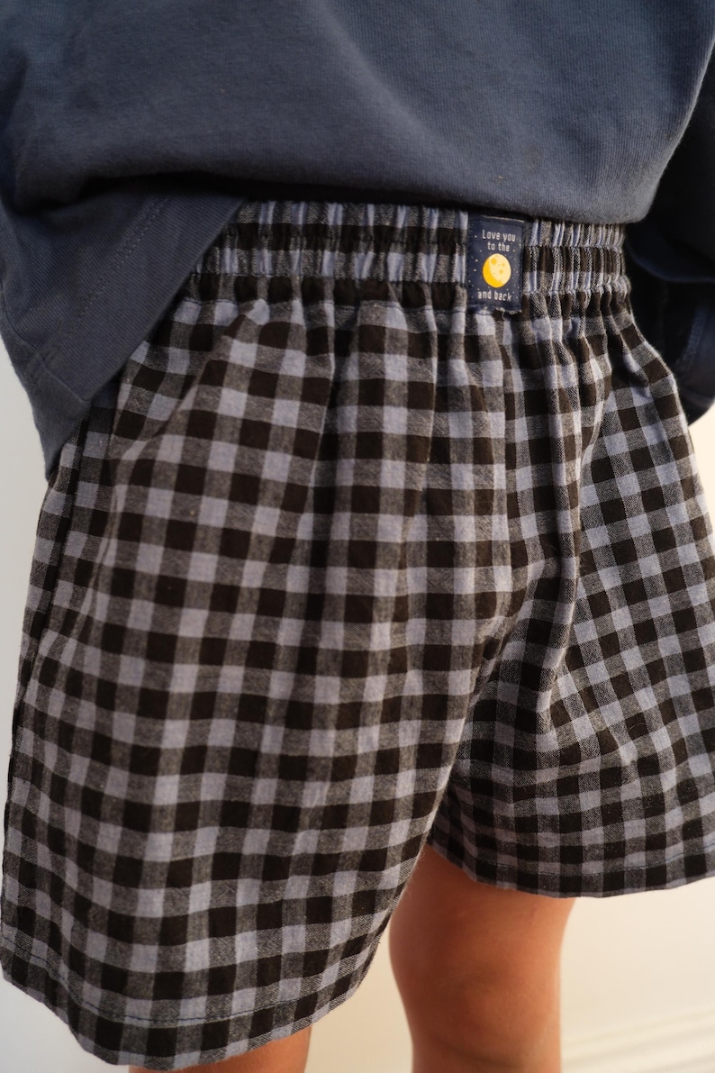 May include: Boxer shorts featuring a black and gray gingham check pattern with an elastic waistband. A small rectangular label on the front center displays a yellow moon graphic and the text "Love you to the moon and back" in white font.