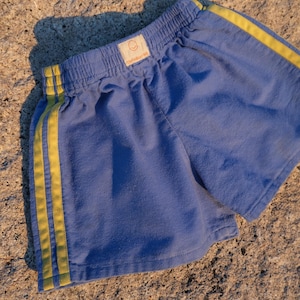 May include: Blue athletic shorts featuring an elastic waistband and two vertical yellow stripes running down each side. A small rectangular label is centered on the front waistband, displaying a smiley face icon and the text The Yellow Hill.