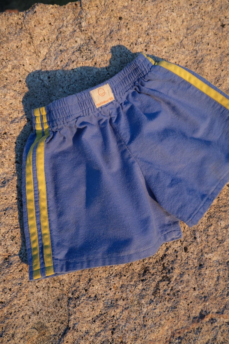 May include: Blue athletic shorts featuring an elastic waistband and two vertical yellow stripes running down each side. A small rectangular label is centered on the front waistband, displaying a smiley face icon and the text The Yellow Hill.