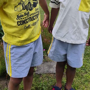 May include: A yellow t-shirt with a black Tonka construction graphic and text, and a white t-shirt with a yellow pocket and truck graphic. Both are paired with light blue shorts featuring yellow side stripes and dark blue and red sandals.