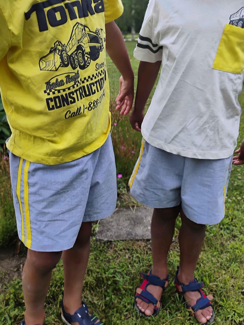 May include: A yellow t-shirt with a black Tonka construction graphic and text, and a white t-shirt with a yellow pocket and truck graphic. Both are paired with light blue shorts featuring yellow side stripes and dark blue and red sandals.