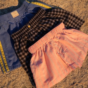 May include: Three pairs of shorts with elastic waistbands. One pair is blue with two yellow side stripes. Another pair features a black and white gingham check pattern. The third pair is light pink with thin white vertical pinstripes.