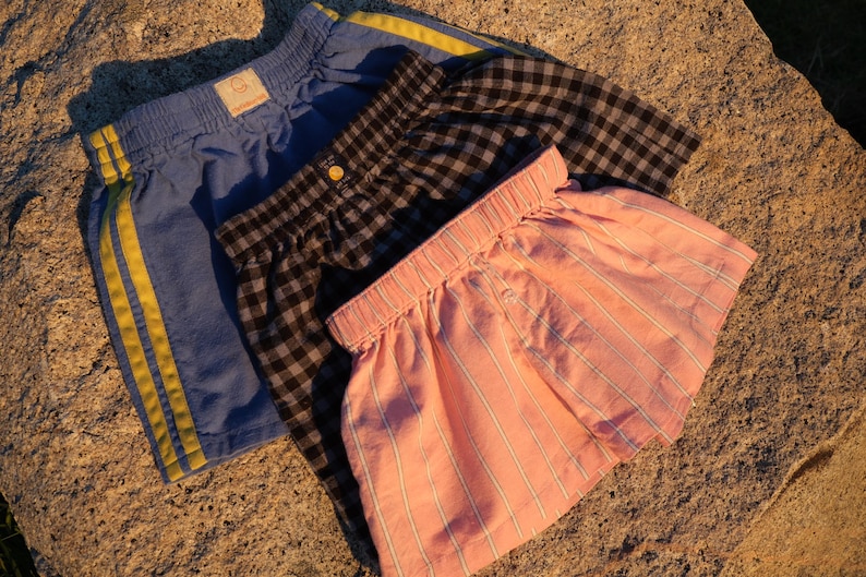 May include: Three pairs of shorts with elastic waistbands. One pair is blue with two yellow side stripes. Another pair features a black and white gingham check pattern. The third pair is light pink with thin white vertical pinstripes.