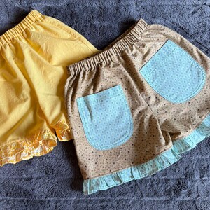May include: Two pairs of elastic-waist shorts. One pair is solid yellow with a matching yellow floral ruffle hem. The second pair is tan with a brown polka dot pattern, featuring two light blue pockets with a white dot print and a light blue ruffle hem.