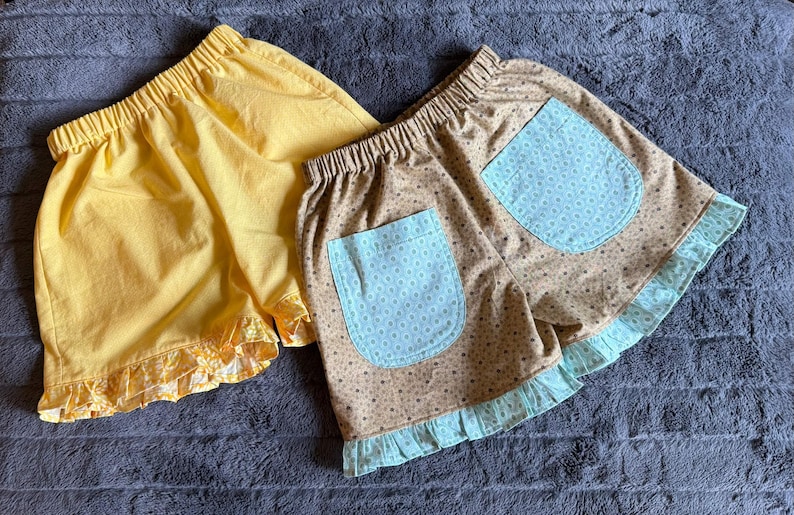 May include: Two pairs of elastic-waist shorts. One pair is solid yellow with a matching yellow floral ruffle hem. The second pair is tan with a brown polka dot pattern, featuring two light blue pockets with a white dot print and a light blue ruffle hem.