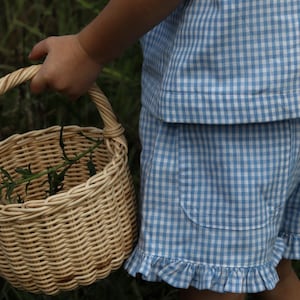 May include: A light blue and white gingham patterned top and shorts set featuring a ruffled hem on the shorts and a patch pocket. The outfit is paired with a round, light-colored woven wicker basket with a handle containing green foliage.