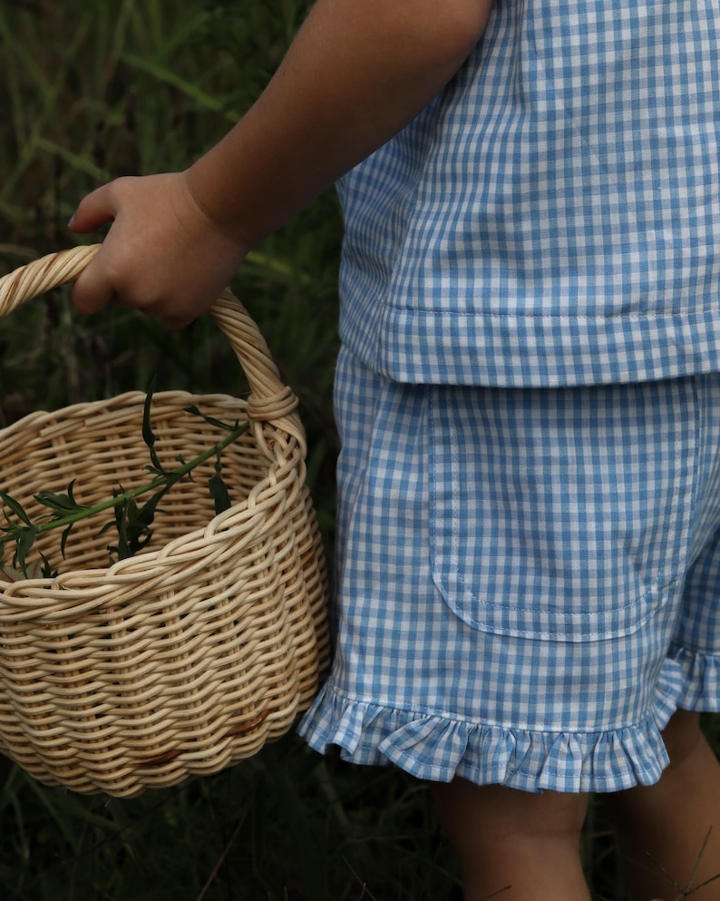 May include: A light blue and white gingham patterned top and shorts set featuring a ruffled hem on the shorts and a patch pocket. The outfit is paired with a round, light-colored woven wicker basket with a handle containing green foliage.