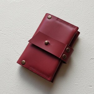 May include: A burgundy leather wallet with a strap closure secured by a brass button. The wallet is rectangular and has a stitched edge. The strap is secured with two additional brass rivets. The wallet is on a white surface.