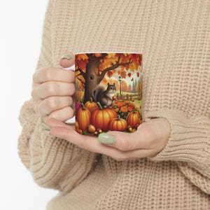 Peut inclure: Mug en céramique blanche avec une scène d'automne : un écureuil, des citrouilles et des feuilles. Le motif comprend un grand arbre, une clôture et un lampadaire, évoquant une ambiance chaleureuse. Parfait pour les boissons chaudes en automne.