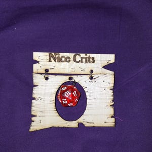 以下が含まれることがあります： 上部に「Nice Crits」の文字が焼き付けられた明るい色の木製ディスプレイプレート。中央に大きな楕円形の切り抜きがある下部の木製パーツから、金属製の丸カンで吊り下げられた半透明の赤い20面ダイスがぶら下がっています。