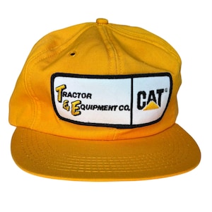 Op de afbeelding: Een gele baseballcap met een platte klep. De pet heeft een rechthoekige patch met de tekst "Tractor & Equipment Co." en het CAT-logo. De hoed is gemaakt van een duurzame stof en heeft een knoop aan de bovenkant.