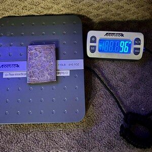 Puede incluir: Un objeto rectangular plateado con un dise&ntilde;o floral adornado se coloca sobre una b&aacute;scula digital gris. La b&aacute;scula muestra un peso de 100,896, y la marca "Accuteck" es visible. La b&aacute;scula tambi&eacute;n muestra "110LB d=0.10Z".