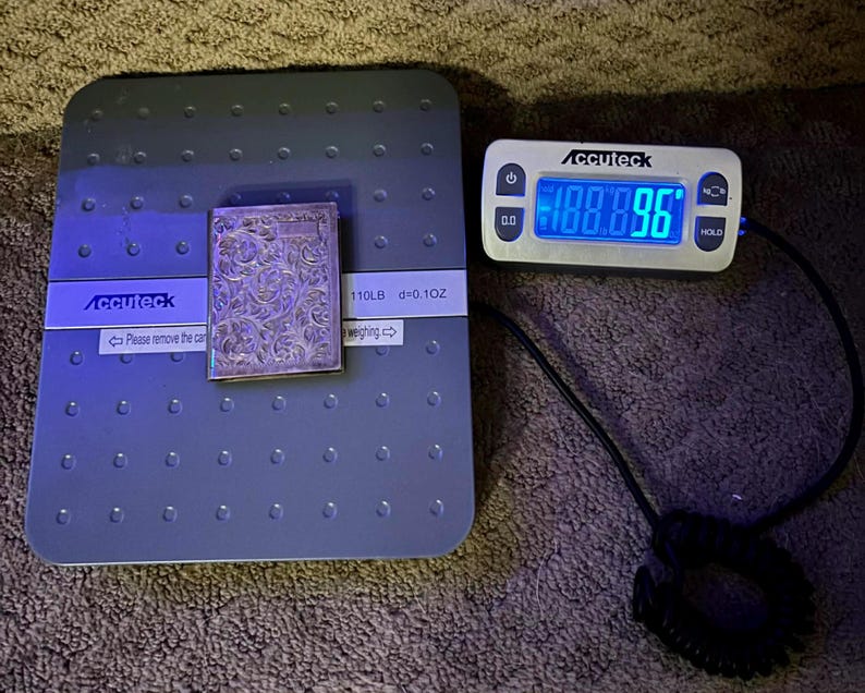 Puede incluir: Un objeto rectangular plateado con un dise&ntilde;o floral adornado se coloca sobre una b&aacute;scula digital gris. La b&aacute;scula muestra un peso de 100,896, y la marca "Accuteck" es visible. La b&aacute;scula tambi&eacute;n muestra "110LB d=0.10Z".