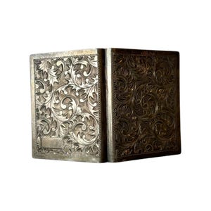 Puede incluir: Una caja rectangular plateada con un patr&oacute;n floral ornamentado grabado en su superficie. La caja est&aacute; abierta, revelando su interior. El intrincado dise&ntilde;o cubre toda la superficie, creando una est&eacute;tica vintage.