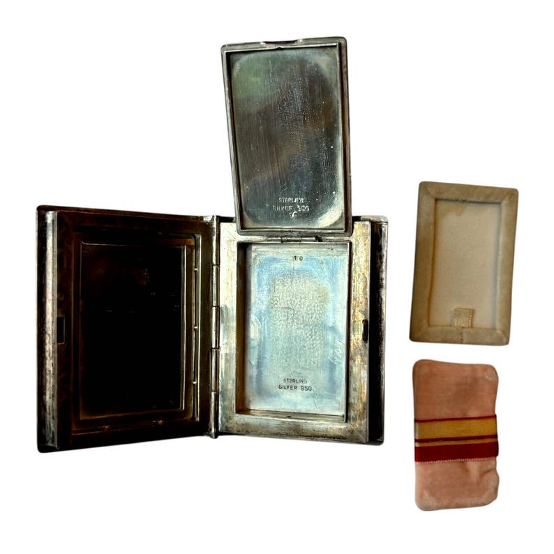Puede incluir: Un compacto de plata de ley antiguo y abierto, con tapa abisagrada, que revela un espejo y compartimentos interiores. El compacto es rectangular con un acabado plateado deslustrado. Tambi&eacute;n hay un peque&ntilde;o marco rectangular y una borla de terciopelo rosa.