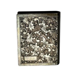 Puede incluir: Un estuche rectangular plateado con intrincados adornos florales. El estuche tiene una tapa con bisagras y un peque&ntilde;o espacio rectangular en blanco cerca de la parte inferior. El grabado detallado cubre toda la superficie, creando una est&eacute;tica vintage.