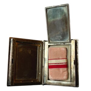Puede incluir: Compacto plateado abierto con forma rectangular. El interior revela una borla de polvo rosa con rayas rojas y blancas. La tapa est&aacute; abierta, revelando un espejo. El compacto est&aacute; marcado "STERLING SILVER 360".