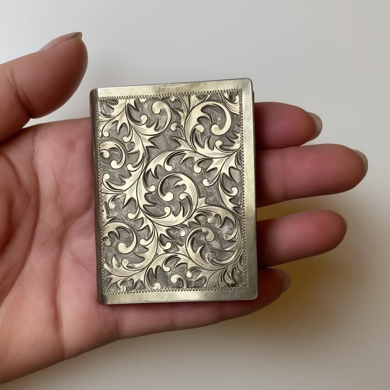 Puede incluir: Un estuche rectangular de metal plateado con un intrincado dise&ntilde;o floral en espiral. El estuche se sostiene en una mano, mostrando el grabado detallado y el contraste entre el metal pulido y el dise&ntilde;o grabado.