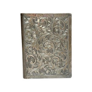 Puede incluir: Un estuche rectangular de metal plateado con un patr&oacute;n floral grabado y ornamentado. El dise&ntilde;o presenta hojas y vides arremolinadas, con un borde texturizado. El estuche parece ser vintage o antiguo.