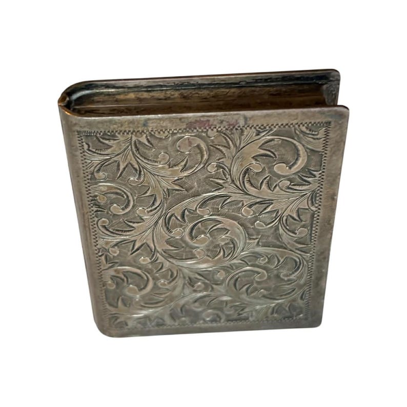 Puede incluir: Una caja rectangular plateada con tapa abisagrada, que se asemeja a un peque&ntilde;o libro. La caja est&aacute; adornada con un intrincado patr&oacute;n floral grabado en toda su superficie. Los bordes de la caja son ligeramente redondeados, lo que sugiere una est&eacute;tica vintage o antigua.