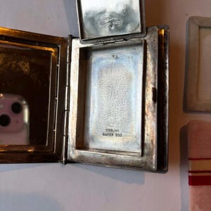 Puede incluir: Un estuche compacto de plata antigua abierto, con tapa abisagrada. El interior presenta un espejo en un lado y un compartimento con el texto "STERLING SILVER 950" en el otro. El estuche es rectangular con esquinas redondeadas.