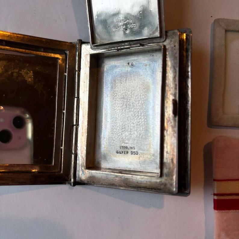 Puede incluir: Un estuche compacto de plata antigua abierto, con tapa abisagrada. El interior presenta un espejo en un lado y un compartimento con el texto "STERLING SILVER 950" en el otro. El estuche es rectangular con esquinas redondeadas.