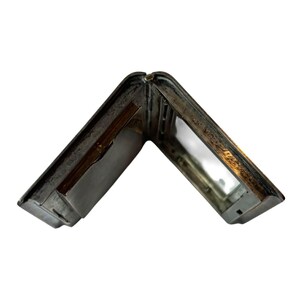 Puede incluir: Un pitillera plateada antigua abierta. La pitillera es rectangular con esquinas redondeadas y una tapa con bisagras. El interior es de color plateado brillante, con un peque&ntilde;o compartimento y una barra con resorte. La pitillera muestra signos de edad y desgaste.