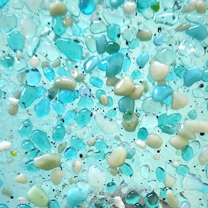 May include: A decorative night light featuring a square design with a mosaic of turquoise, light green, and white glass pebbles. The night light has a white base and is held in a hand.