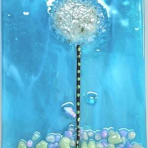 May include: A blue glass night light with a white dandelion design and green and purple glass pebbles at the bottom. The night light has a white on/off switch.