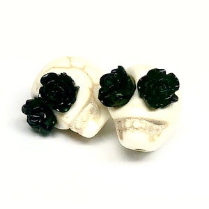 May include: Two white skull beads with black rose accents. The beads are small and have a detailed skull design.