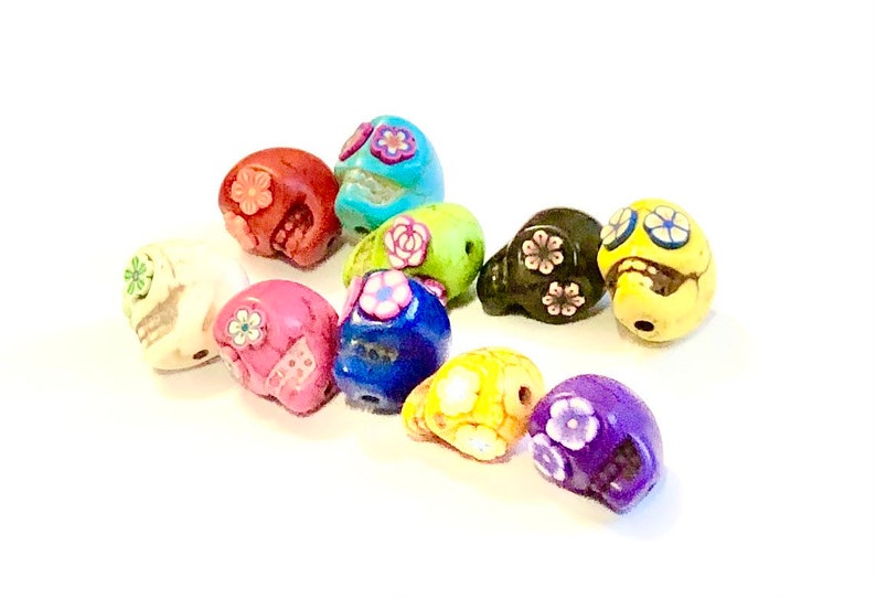 May include: A collection of 12 colorful skull beads with floral designs. The beads are made of ceramic and are approximately 1 inch in size. They are perfect for making jewelry or other crafts.