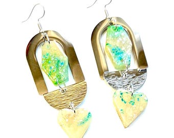May include: A pair of dangle earrings with a unique design. Each earring features a gold-colored arch, a textured silver-colored half-circle, and a heart-shaped charm. The earrings have a light yellow and green speckled stone in the center.
