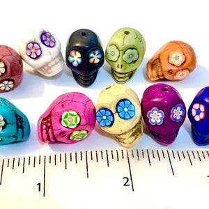 May include: Ten colorful sugar skull beads with floral designs. The beads are approximately 1 inch in diameter and are arranged on a ruler.